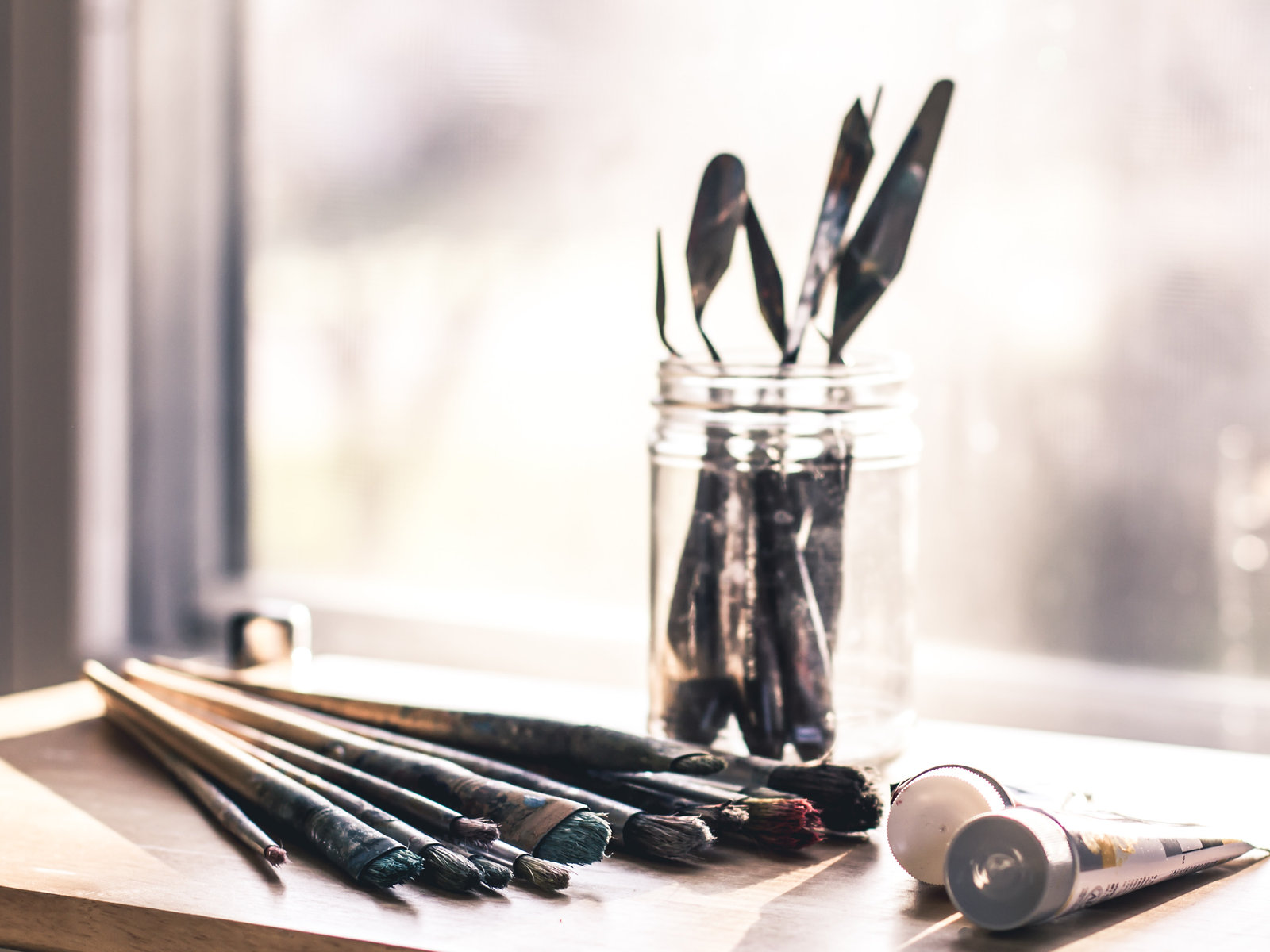 A jar of brushes and paint tubes on a sunny studio windowsill