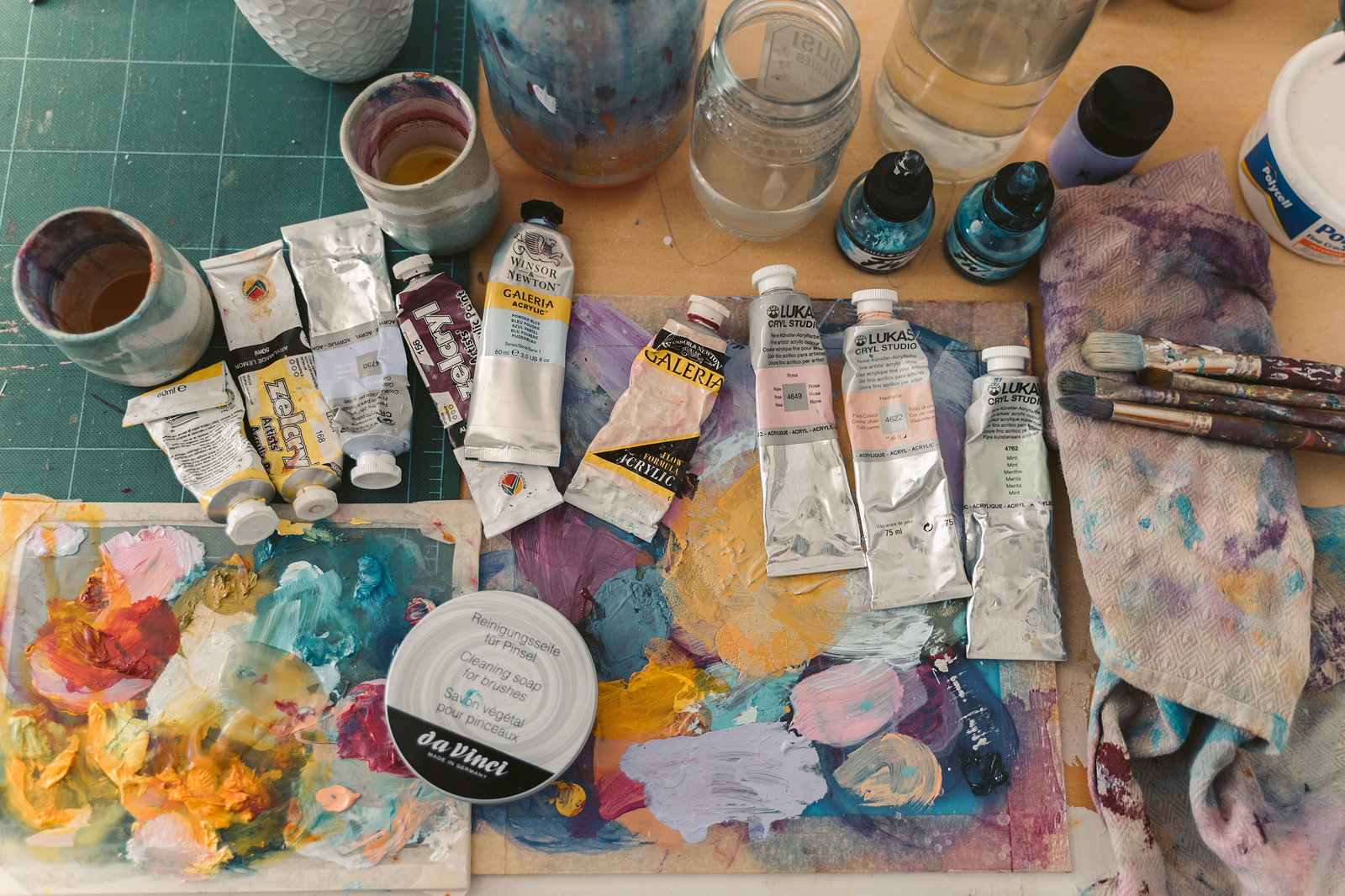 Tubes of paint, brushes and a palette of soft pink, yellow and teal colour on a studio worktable