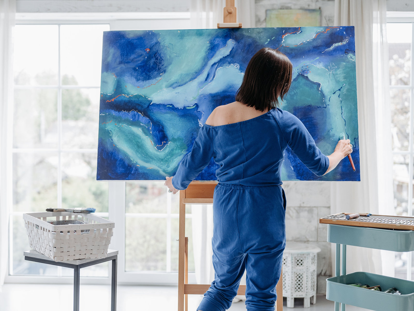 A painter in a blue jumpsuit working on a large teal and blue abstract canvas on a wooden easel in a bright, sunlit studio