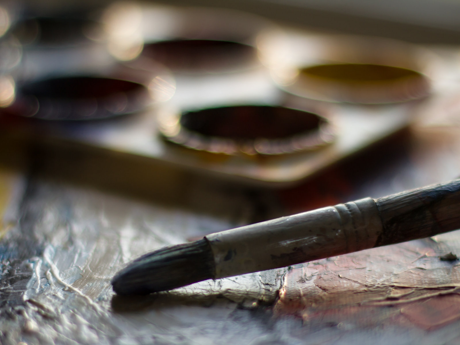 A paintbrush resting on a worn palette table beside a tray of paint wells